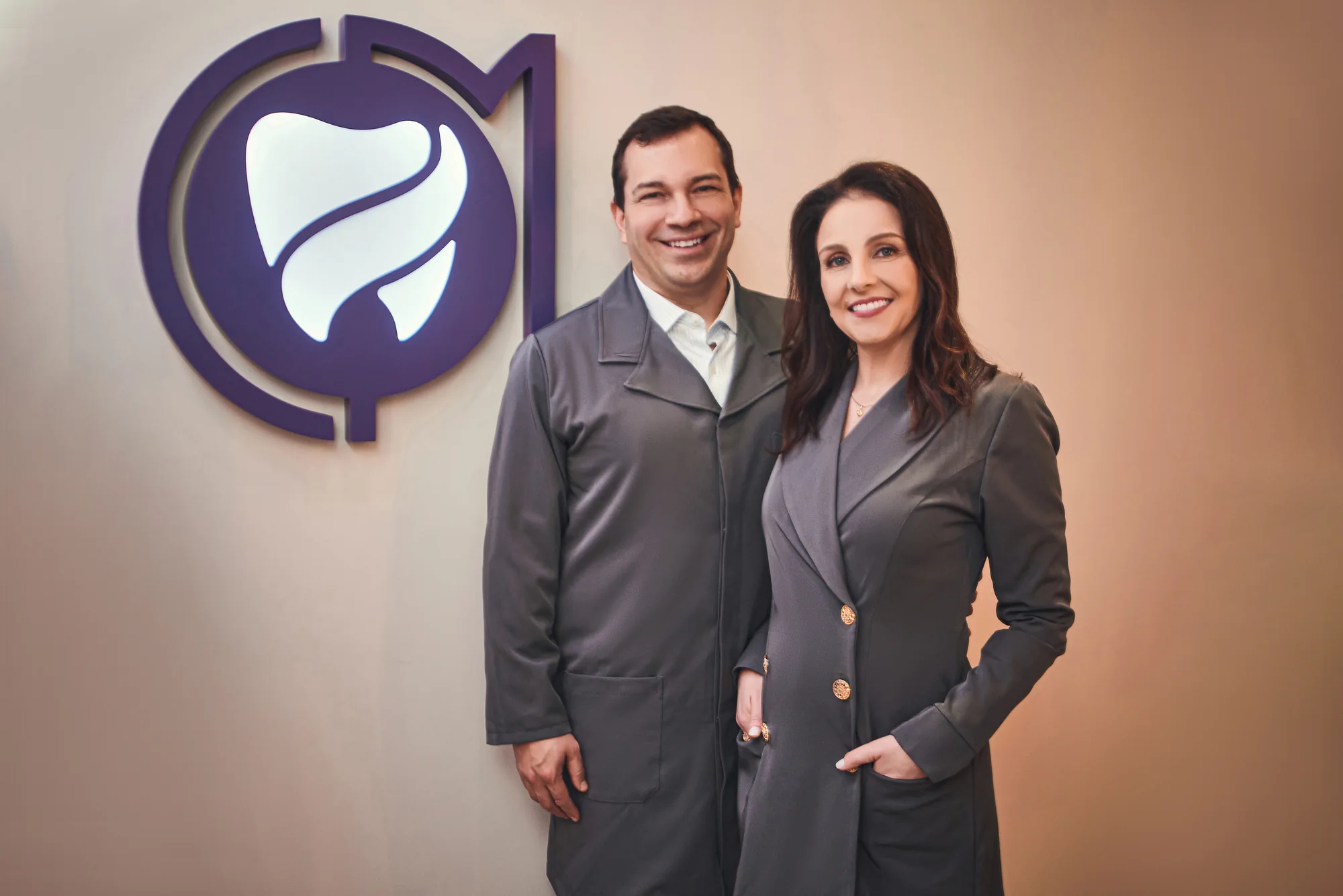Charles Cavalheiro e Lisandra Mozzaquatro juntos, em jalecos cinza, diante da marca da clínica.